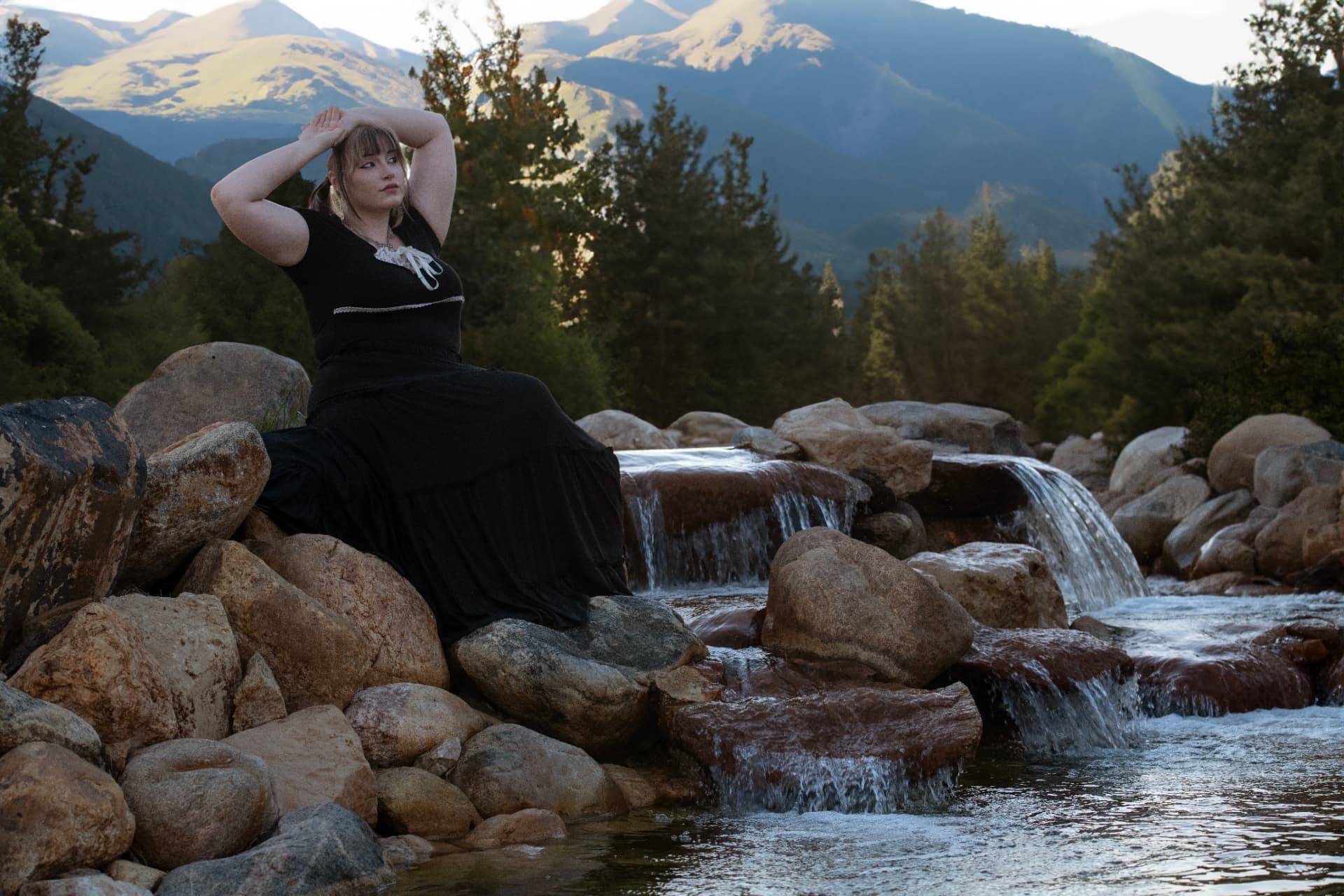 Senior in a long black dress seated on boulders beside a mountain waterfall at dusk