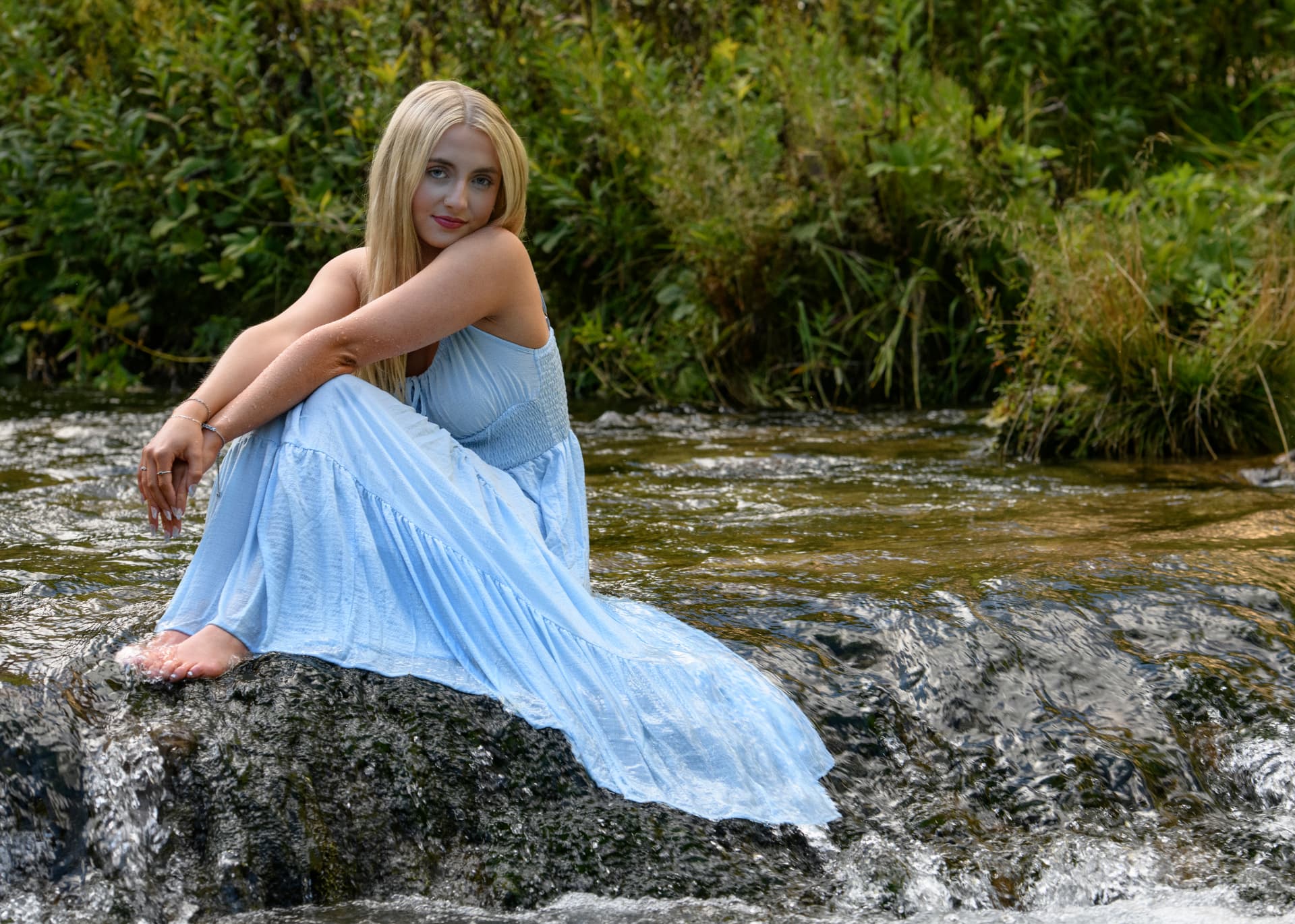 High school senior in a flowing light-blue dress seated on a mossy creek rock, surrounded by green foliage and moving water