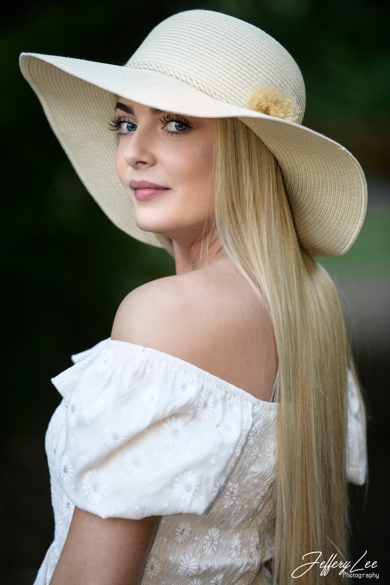 High school senior in a wide-brimmed sun hat and white off-shoulder dress against soft green foliage
