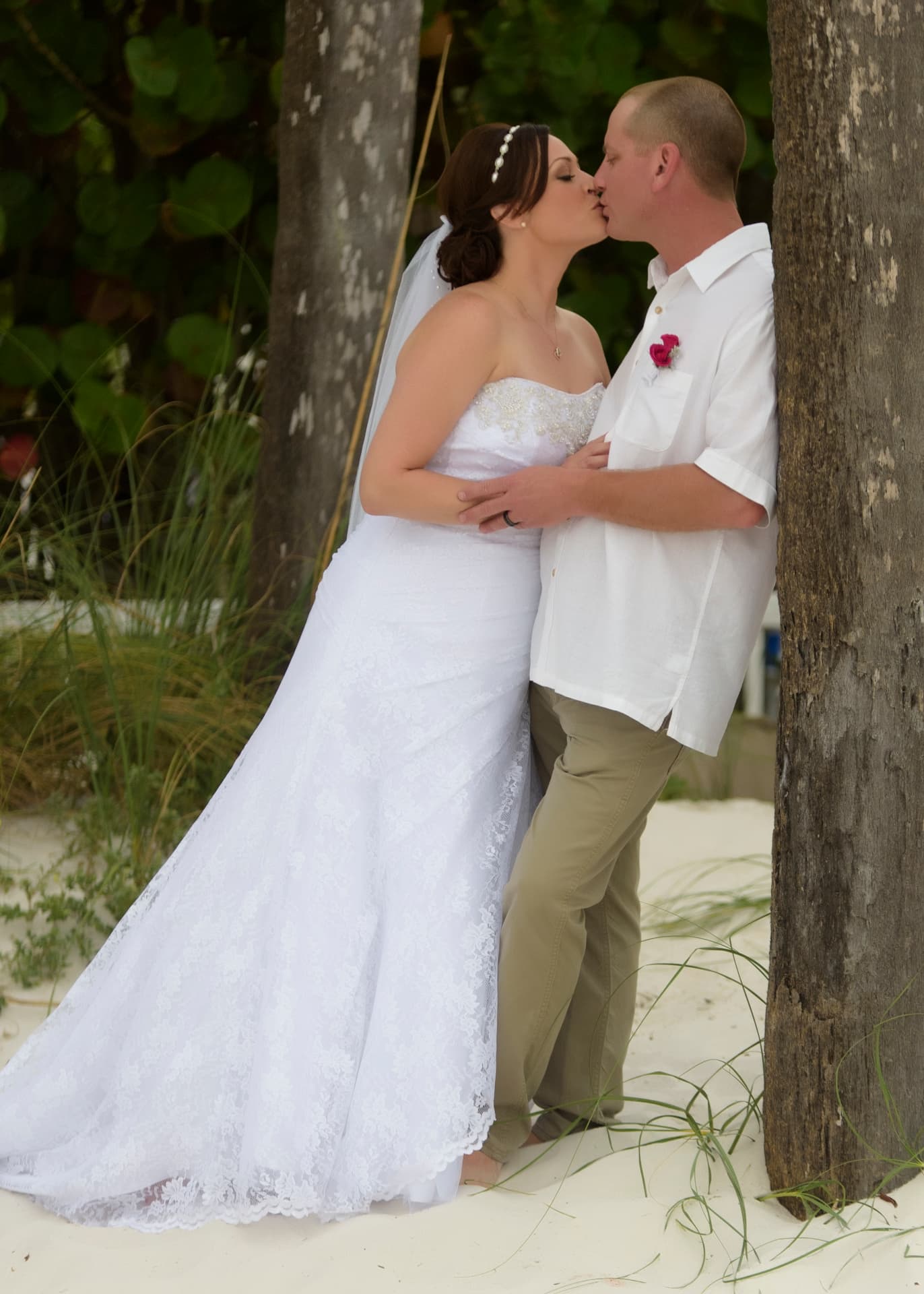 Bride and groom kiss while leaning against a palm tree on a sandy beach, surrounded by tropical greenery