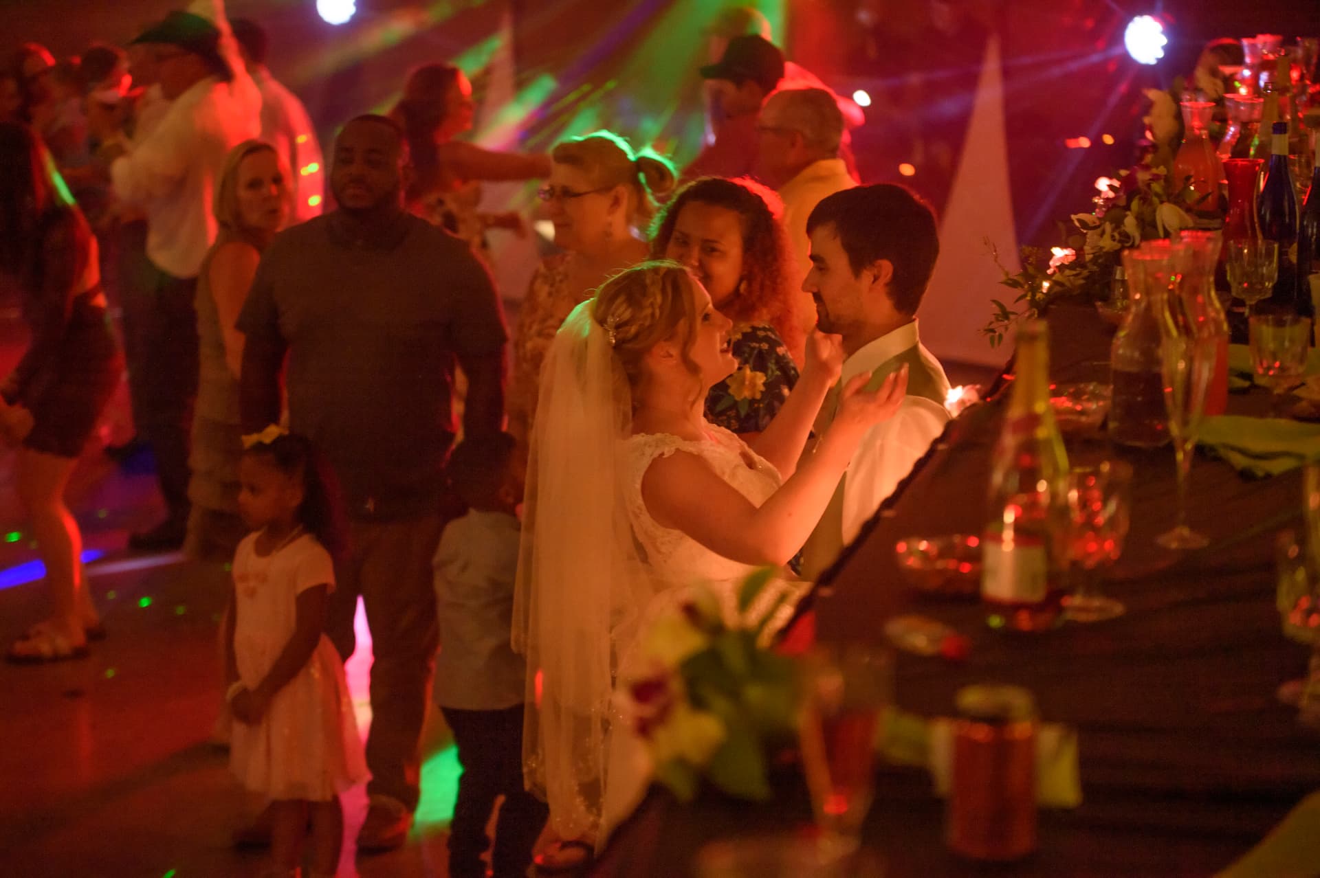 Bride and groom dancing together at a warm, colorful wedding reception surrounded by guests and party lights