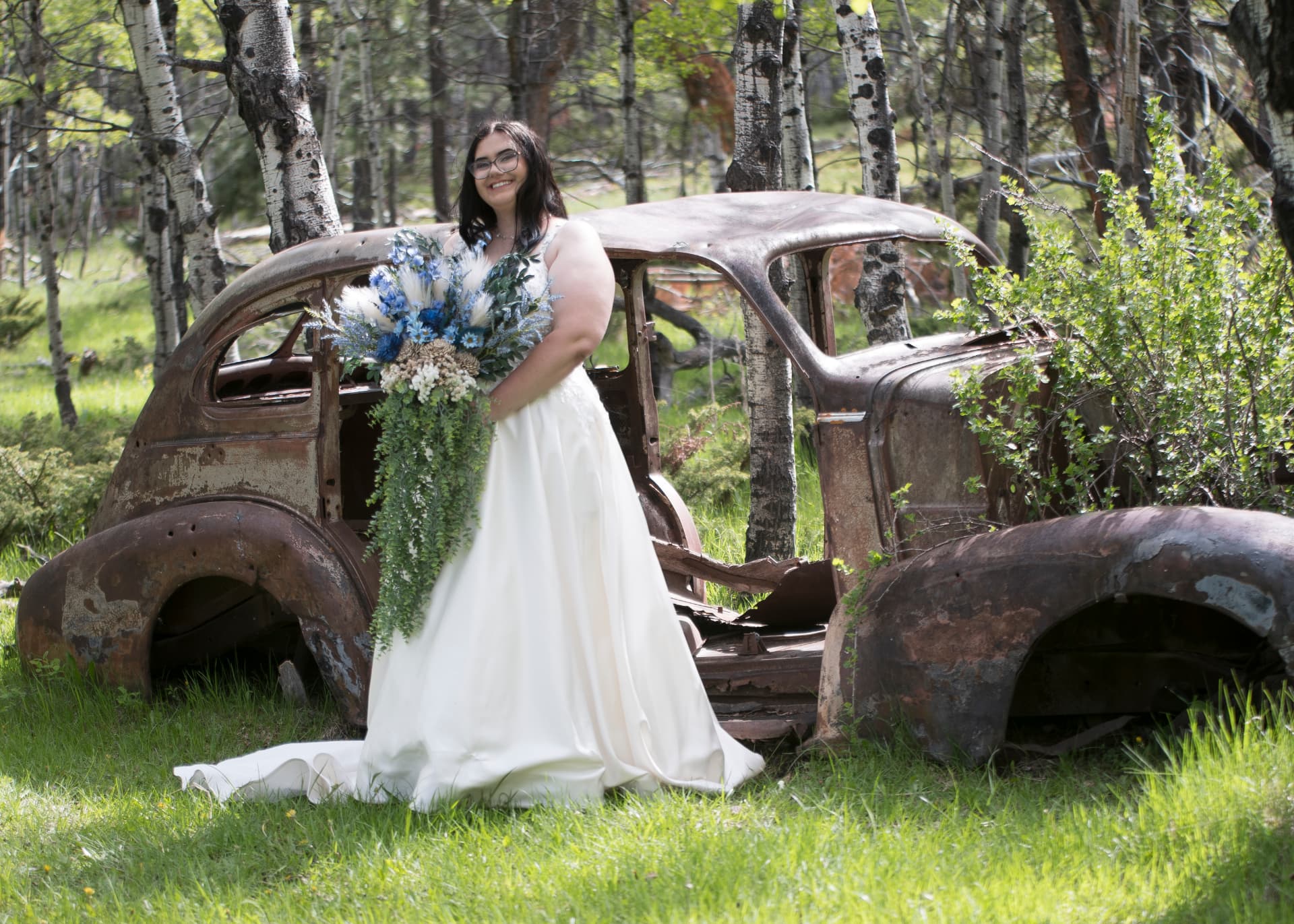 Bride in a flowing white gown holding a blue-and-white bouquet beside a rusted vintage car in an aspen grove