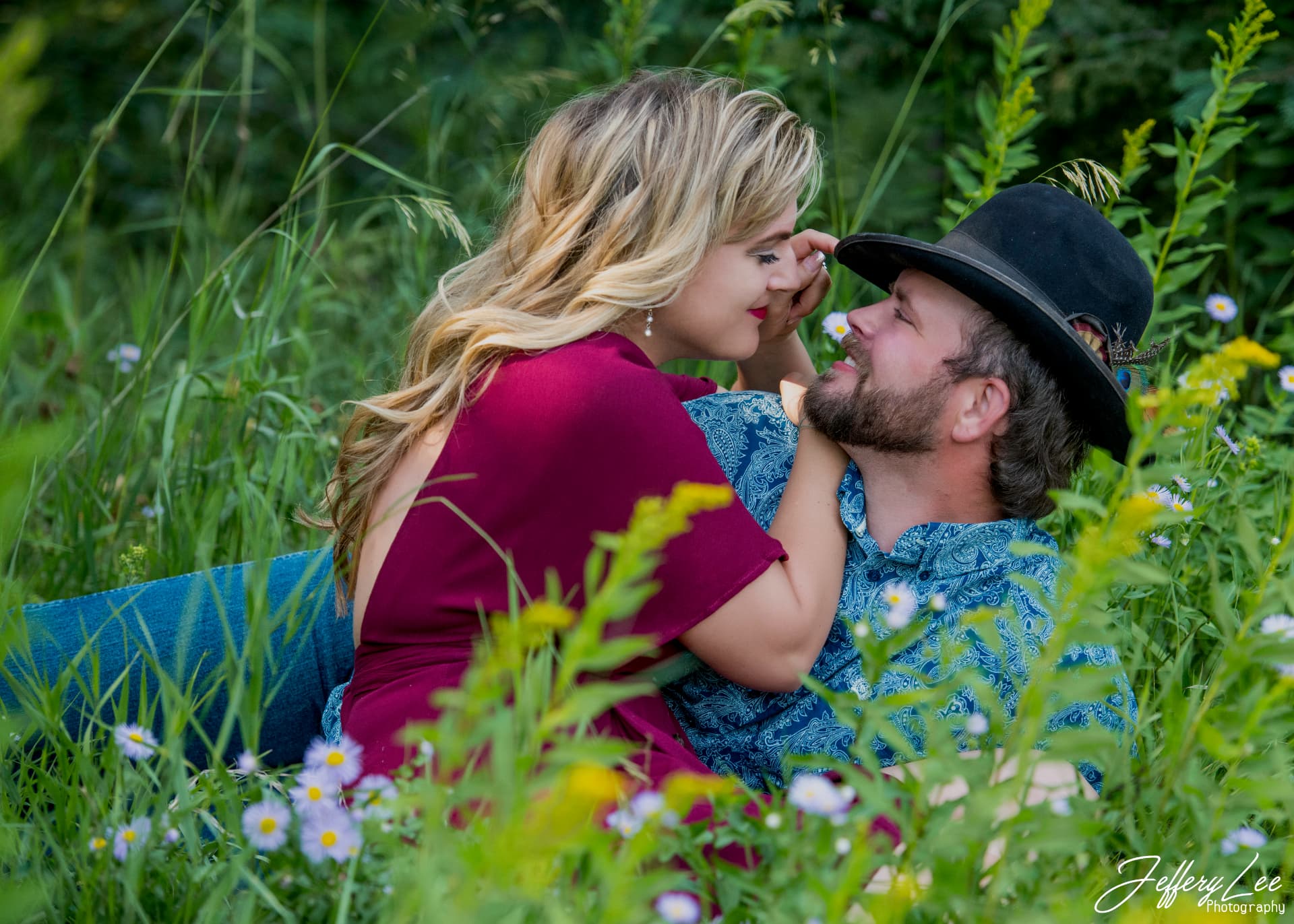 Couple embracing in tall grass and wildflowers, the woman in a deep red dress and the man in a hat